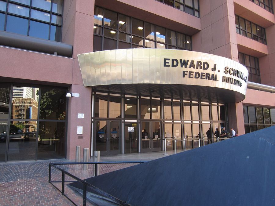 Archive: West entrance of the Edward J. Schwartz Federal Building, San Diego — gold canopy signage and glass entry. Another Believer, License CC BY-SA 4.0. Not the 25 September 2026 verdict day, not day-of Apple or Taction lawyers, and not an iPhone or Apple Watch still.